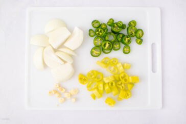 A white cutting board with chopped vegetables: sliced white onion, green jalapeño rounds, yellow bell pepper pieces, and small chopped garlic cloves, all arranged in separate sections.