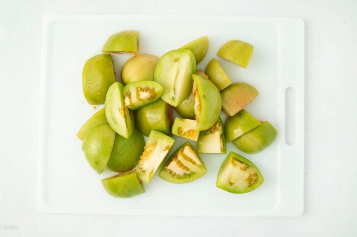 Chopped green tomatoes are arranged on a white cutting board, viewed from above on a white background.