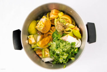 A metal pot filled with chopped green tomatoes, onions, cilantro, and a variety of ground spices, viewed from above against a white background.