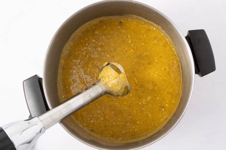 A pot filled with thick yellow soup being blended with an immersion blender on a white surface.