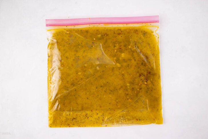 A clear ziplock bag filled with yellowish marinade containing herbs and spices, placed on a plain white background.