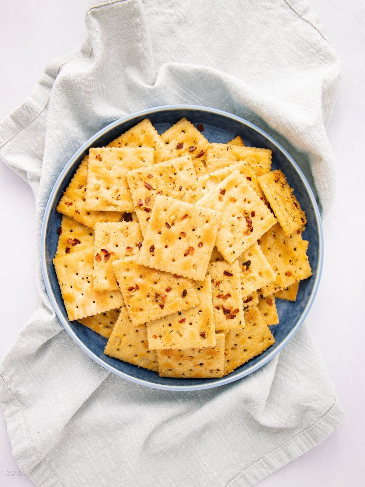 A blue bowl filled with square, seasoned crackers sits on a light gray cloth, viewed from above. The crackers have a golden color and are sprinkled with herbs or spices.
