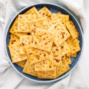 A blue plate filled with square crackers sprinkled with herbs and seasonings, placed on a light-colored cloth.