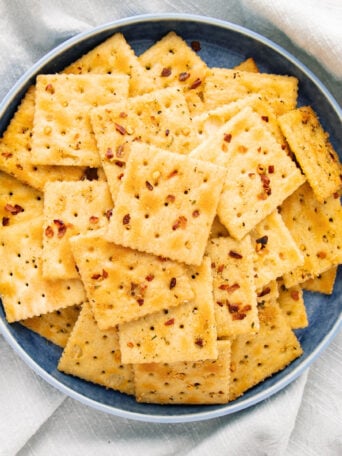 A blue plate filled with square crackers sprinkled with herbs and seasonings, placed on a light-colored cloth.
