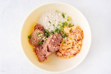A mixing bowl containing ground meat, chopped green onions, shredded cheese with red bits, and flour, all separated into sections, ready to be mixed together.