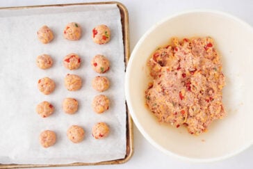 A bowl of raw meat mixture with chopped vegetables sits next to a baking tray lined with parchment paper, holding evenly spaced, uncooked meatballs.