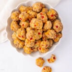 A white bowl filled with small, round sausage balls containing visible bits of red and green peppers, set on a white surface with a white cloth partially visible in the corner.