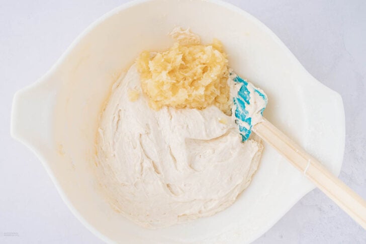 A white mixing bowl with thick batter and a mound of crushed pineapple on top, next to a spatula with a blue tip.