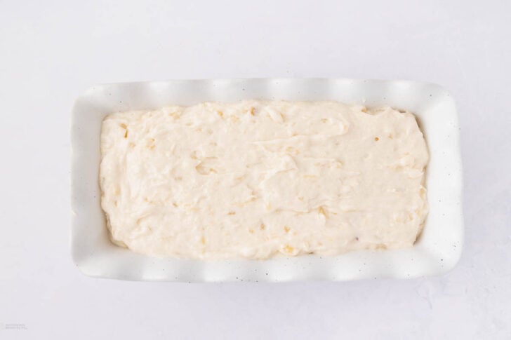 A white rectangular baking dish filled with unbaked, creamy banana bread batter, spread evenly and ready to be baked, on a white surface.
