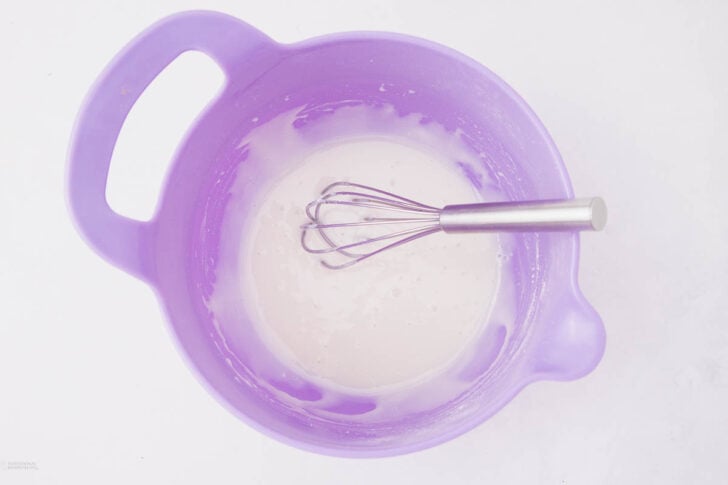 A purple mixing bowl with a metal whisk inside, containing a smooth white batter or mixture, set against a white background.
