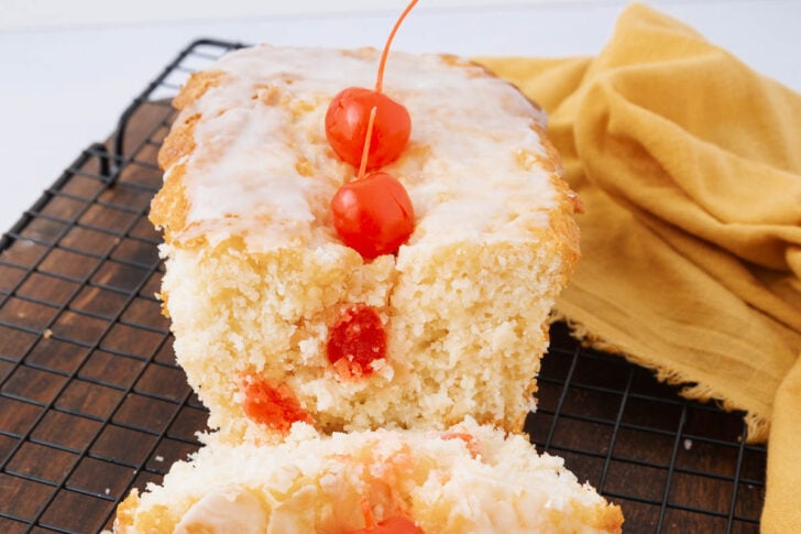 A loaf of glazed cherry bread with two maraschino cherries on top and visible cherry pieces inside, resting on a cooling rack next to a yellow cloth.