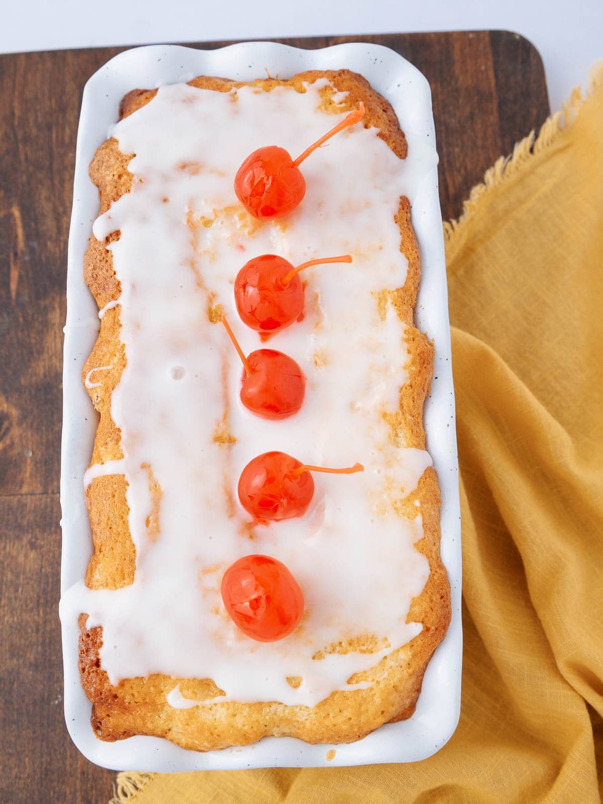 A rectangular loaf cake with white icing drizzled on top, garnished with five red cherries in a row, sits on a wooden surface next to a yellow cloth.