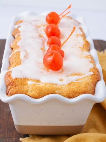 A rectangular loaf cake with white icing and topped with three red maraschino cherries sits in a white ceramic dish on a wooden surface next to a yellow cloth.