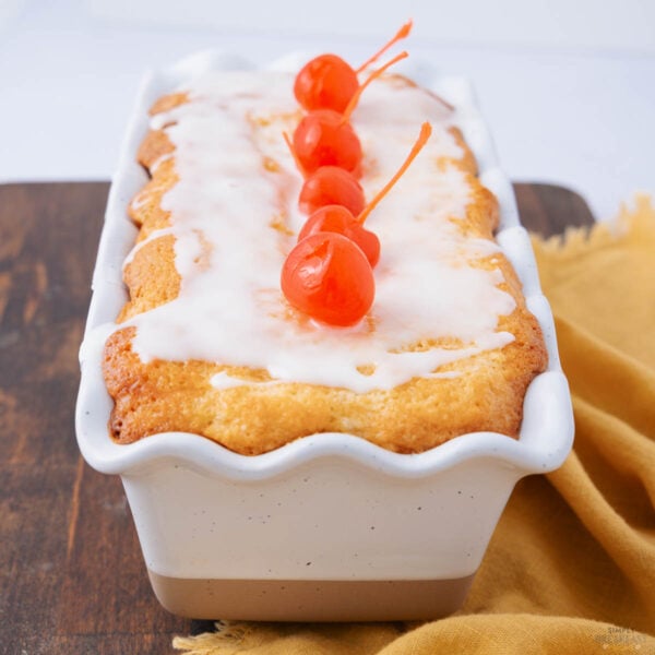 A rectangular loaf cake with white icing and topped with three red maraschino cherries sits in a white ceramic dish on a wooden surface next to a yellow cloth.