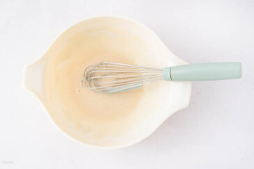 A light-colored mixing bowl with a pale green-handled whisk inside, containing a small amount of light tan liquid batter. The bowl sits on a white surface.