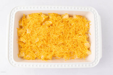 A white rectangular baking dish filled with uncooked casserole, topped with a generous layer of shredded cheddar cheese, on a white background.
