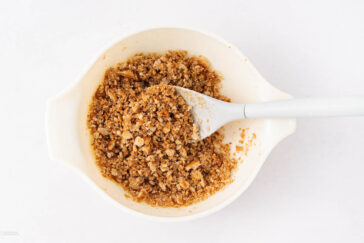 A white mixing bowl containing a crumbly mixture of crushed graham crackers, sugar, and melted butter, with a white spatula resting inside.