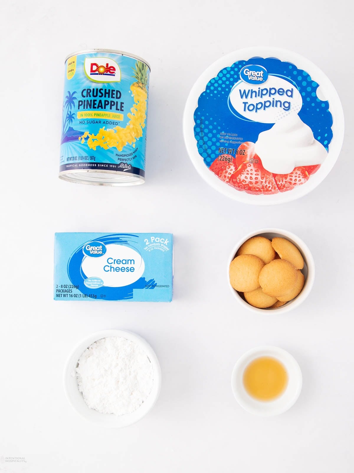 Five ingredients arranged on a white surface: a can of crushed pineapple, a tub of whipped topping, a box of cream cheese, a small bowl of vanilla wafers, a bowl of powdered sugar, and a small bowl of vanilla extract.