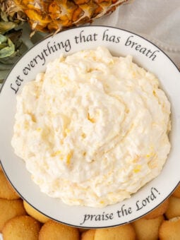 A bowl of creamy pineapple dip surrounded by vanilla wafer cookies sits on a plate with the words "Let everything that has breath praise the Lord!" written around the rim. A pineapple top is visible nearby.