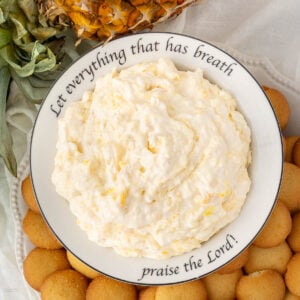 A bowl of creamy pineapple dip surrounded by vanilla wafer cookies sits on a plate with the words "Let everything that has breath praise the Lord!" written around the rim. A pineapple top is visible nearby.