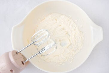A hand mixer with two beaters is blending creamy white batter in a large white mixing bowl on a light surface.