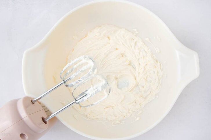 A hand mixer with two beaters is blending creamy white batter in a large white mixing bowl on a light surface.