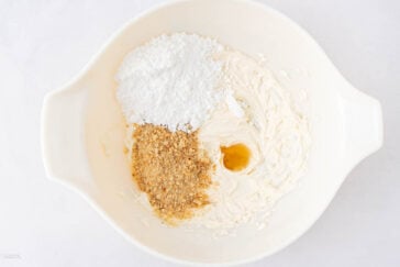 A white mixing bowl containing partially mixed cream cheese, powdered sugar, crushed graham crackers, and a small amount of vanilla extract on a white surface.