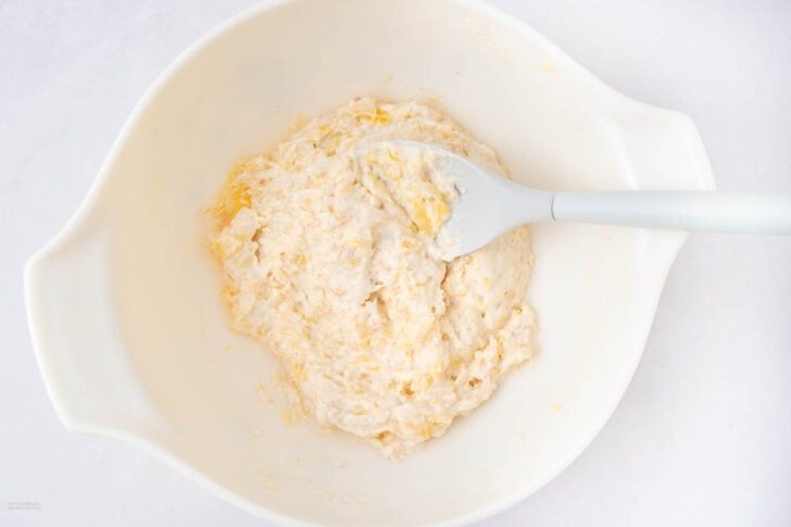 A white mixing bowl with pale, sticky dough being stirred by a white spatula, all set against a white background.