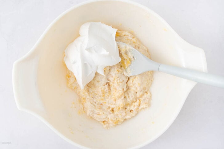 A white bowl containing a partially mixed batter with a dollop of whipped cream on top, and a white spatula resting inside the bowl.