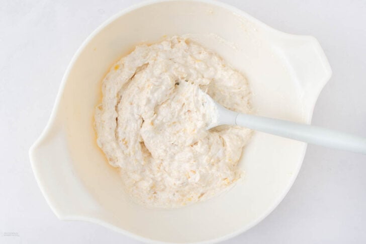 A white mixing bowl containing a thick, creamy dough with a white spatula resting inside, all placed on a white surface.