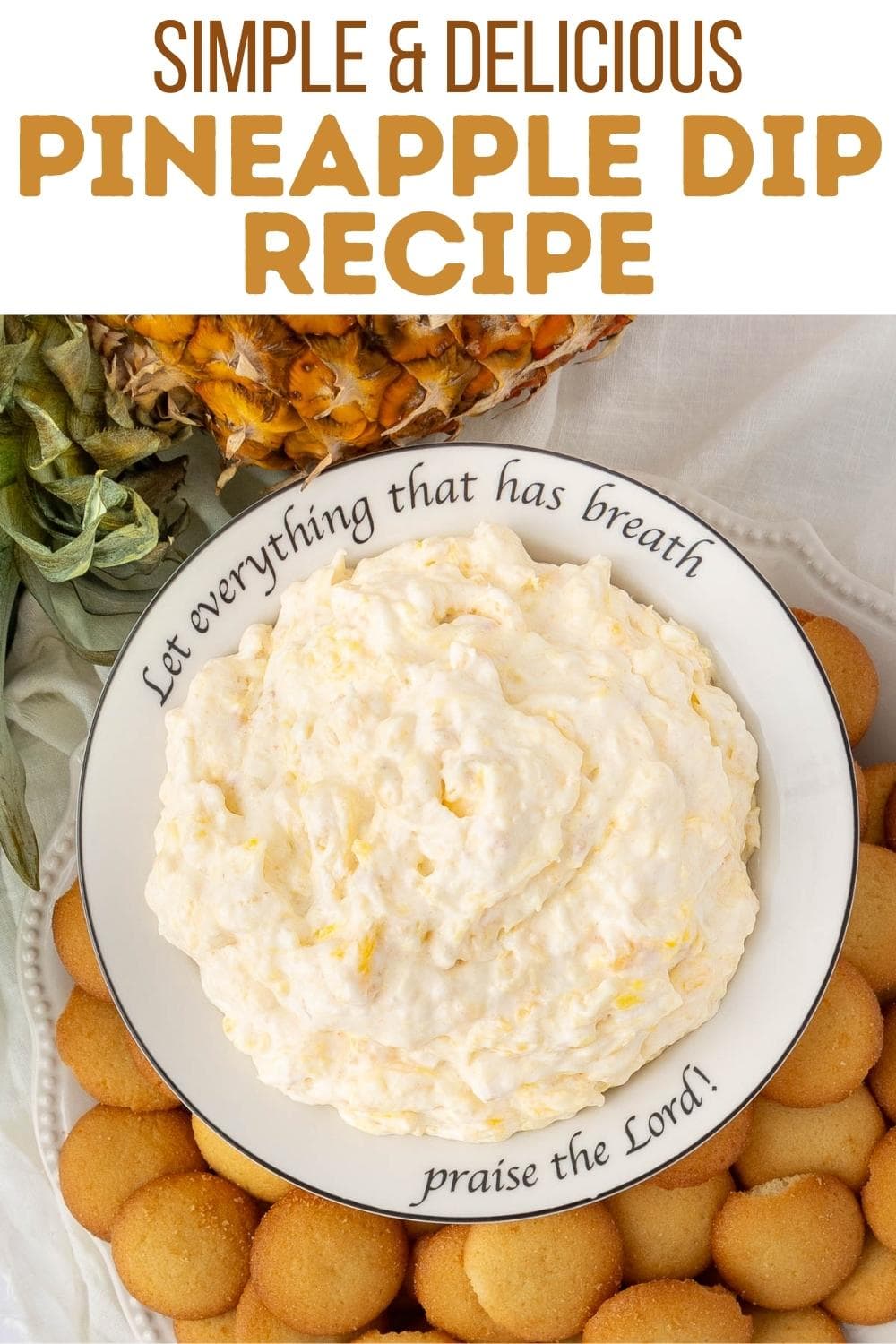 A bowl of creamy pineapple dip sits on a plate surrounded by vanilla wafers, with a pineapple in the background. The plate’s rim reads, “Let everything that has breath praise the Lord!”.