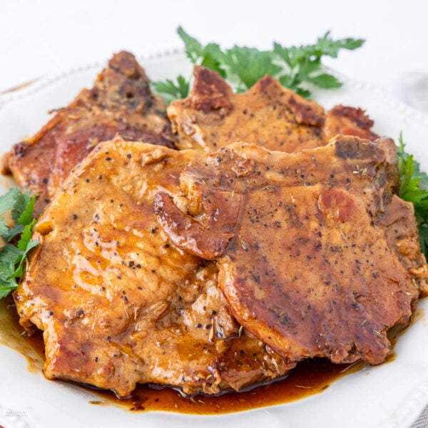 Three seasoned and glazed pork chops are arranged on a white plate, garnished with fresh parsley. The meat appears juicy and tender, coated in a rich brown sauce with visible herbs and spices.