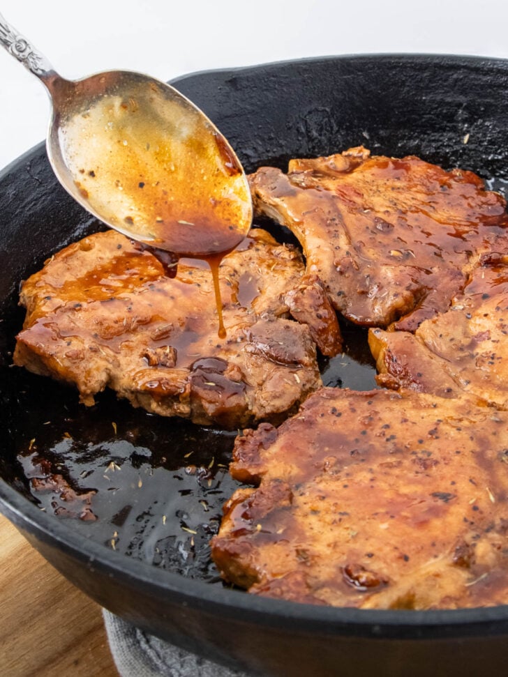 A spoon drizzles sauce over cooked pork chops in a black cast iron skillet, with a glossy brown glaze covering the meat.