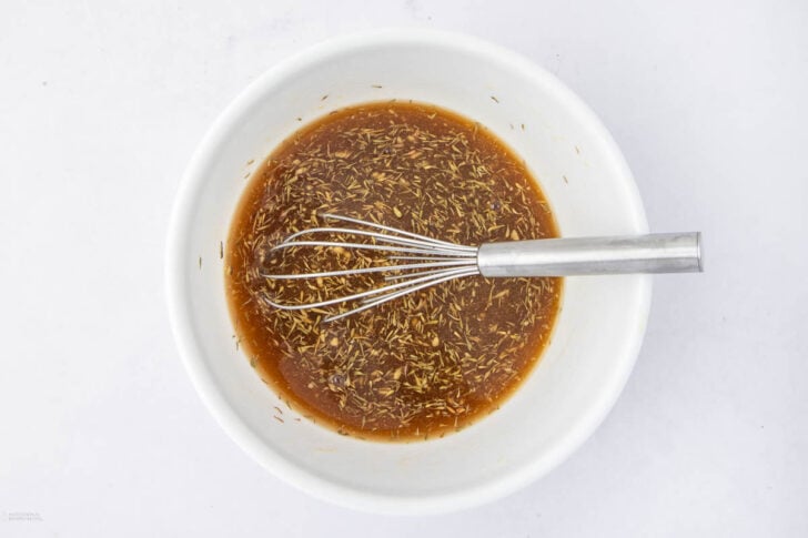 A metal whisk rests in a white bowl filled with a brown liquid marinade mixed with visible dried herbs, set on a white surface.