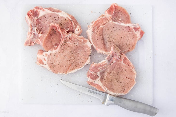 Four raw bone-in pork chops seasoned with black pepper on a white cutting board, with a knife placed below the chops.