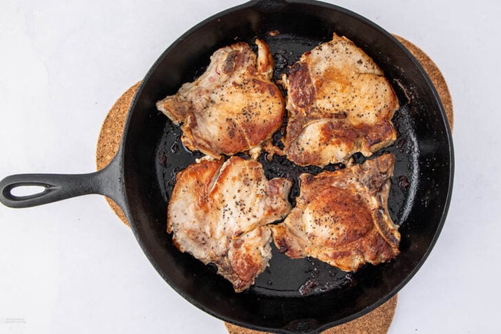 A cast iron skillet containing four cooked, bone-in pork chops seasoned with pepper, placed on a round cork trivet against a white background.