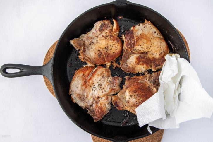 Four seasoned, cooked chicken thighs in a black cast iron skillet, with a white paper towel placed under one piece. The skillet rests on a round cork trivet, set against a white background.