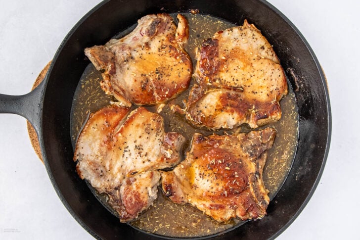 A black skillet containing four cooked, seasoned pork chops with a light brown sauce, viewed from above on a white background.
