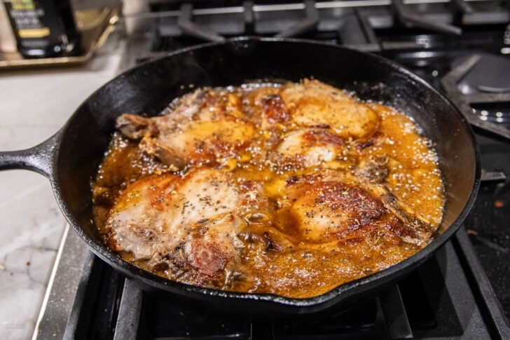 Bone-in pork chops are being pan-fried in a cast iron skillet, sizzling in bubbling oil or sauce on a stovetop. The pork chops have a golden-brown, crispy exterior.