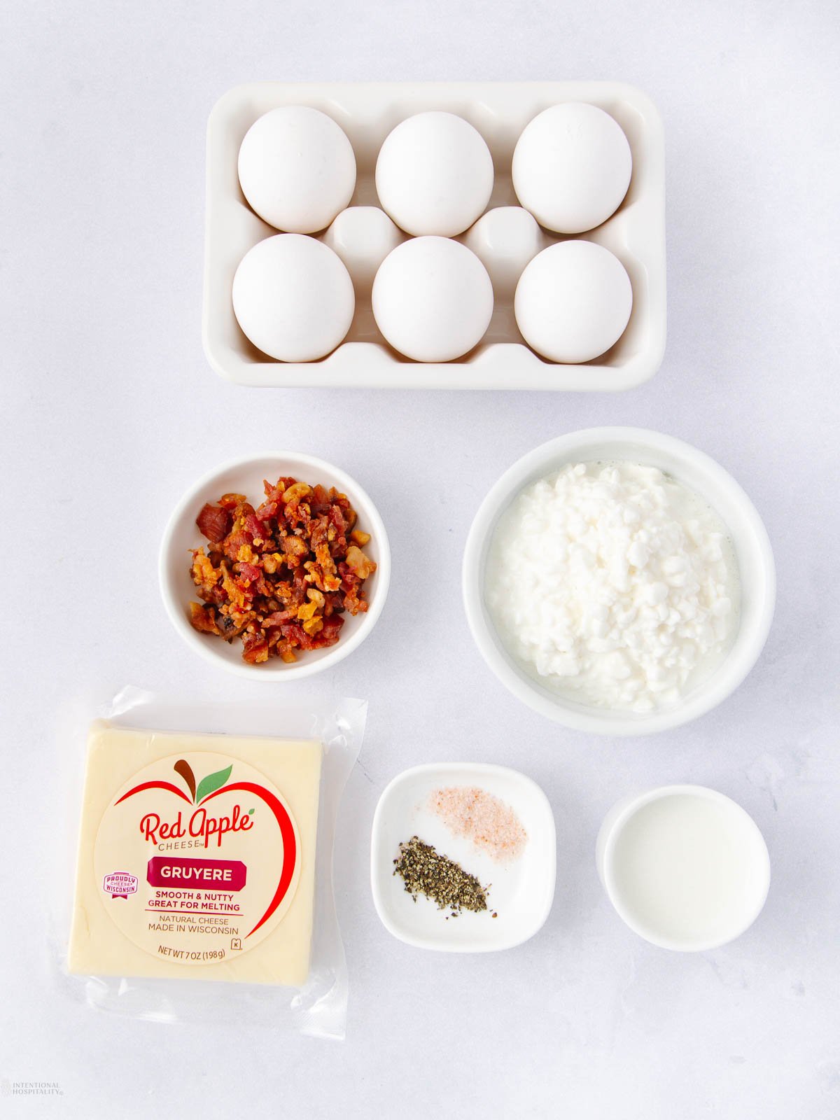 A flat lay of ingredients including six eggs, chopped cooked bacon, cottage cheese, Gruyere cheese slices, a small bowl of milk, a dish of salt and pepper, all arranged on a white surface.