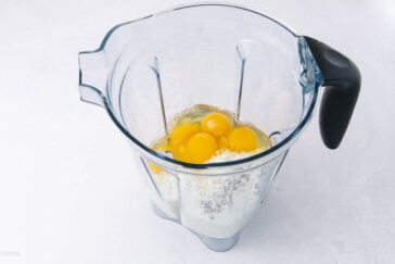 A blender pitcher containing raw eggs, flour, and milk, ready to be mixed, sits on a white surface.