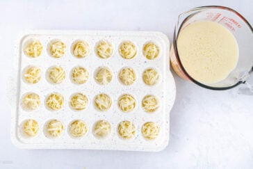 A white muffin tray with shredded cheese in each cup sits beside a glass measuring cup filled with a pale yellow liquid mixture on a white surface.