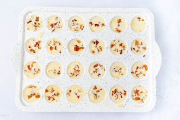 A white, speckled silicone mold tray with 24 round cavities filled with a creamy batter, each topped with small pieces of chopped bacon. The tray is set on a white surface.