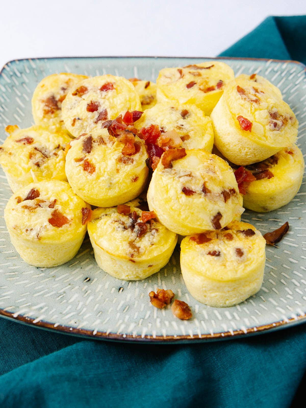 A plate of mini egg muffins topped with small bacon pieces, arranged in a pile on a square, speckled dish, set on a dark teal cloth.