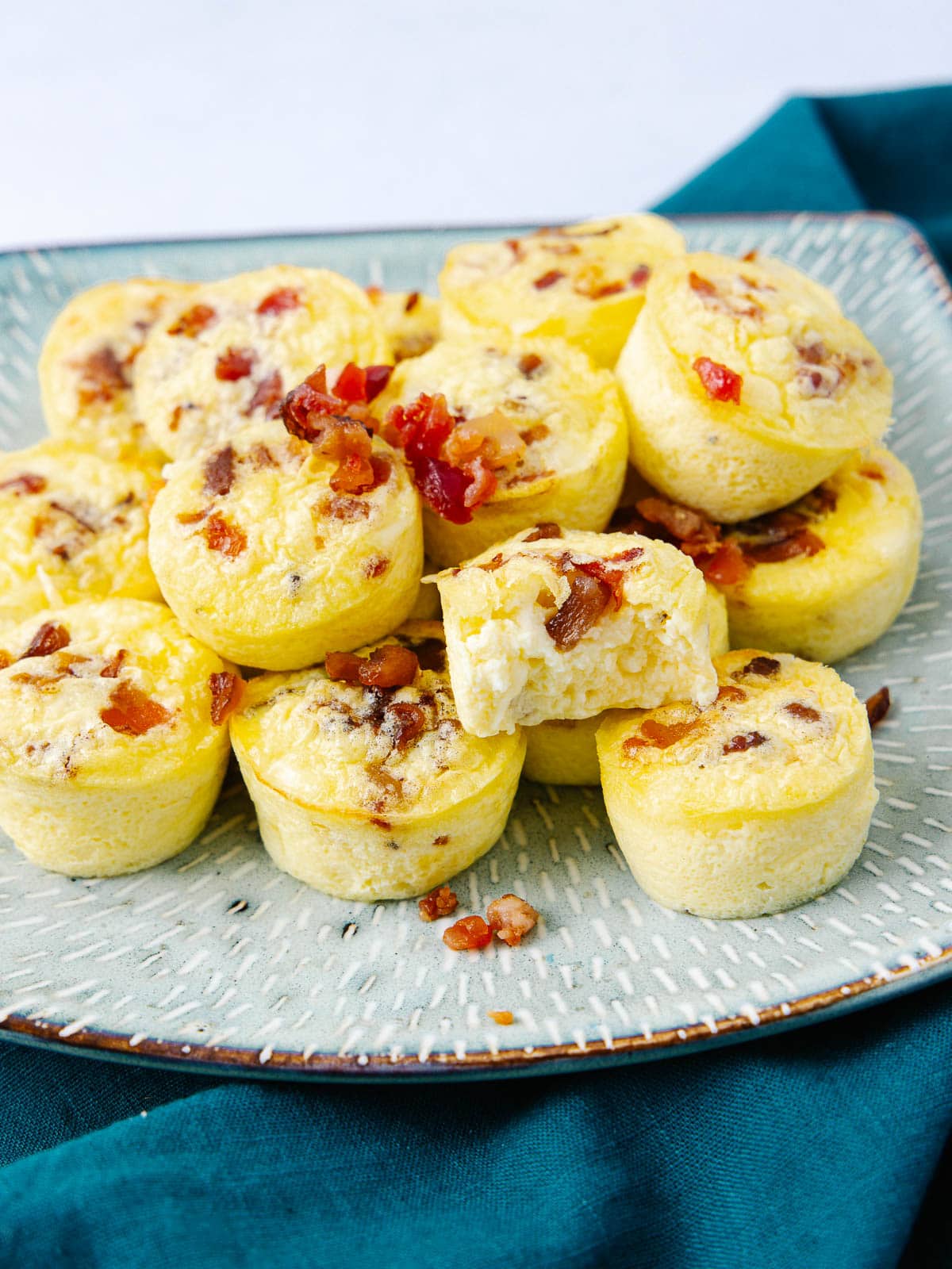 A plate of mini egg bites with bacon pieces on top, arranged on a speckled ceramic plate over a teal cloth. One bite is missing a piece, showing the fluffy interior.