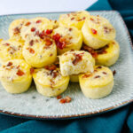 A plate of mini egg bites topped with bits of bacon is shown on a patterned plate. One egg bite is cut open, revealing its fluffy interior. The plate rests on a dark teal cloth.