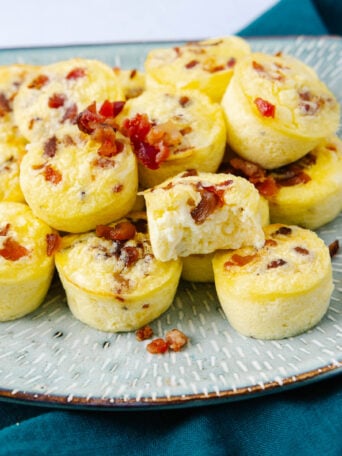 A plate of mini egg bites topped with bits of bacon is shown on a patterned plate. One egg bite is cut open, revealing its fluffy interior. The plate rests on a dark teal cloth.