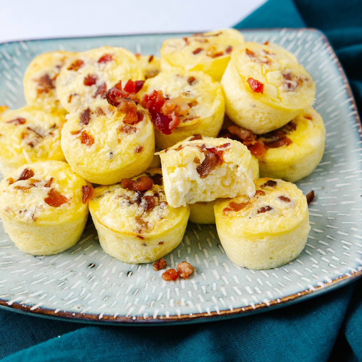A plate of mini egg bites topped with bits of bacon is shown on a patterned plate.
