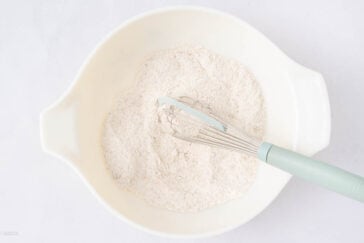 A metal whisk with a light blue handle rests in a white mixing bowl filled with dry flour mixture on a white surface.