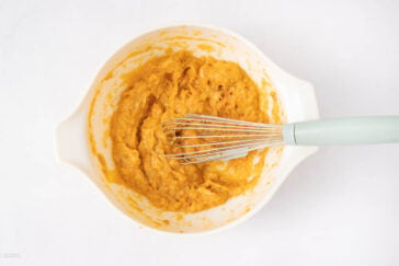 A white mixing bowl filled with a thick, orange batter and a metal whisk with a light blue handle resting inside, all on a white background.
